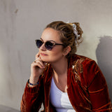 Frau mit hochgesteckten Haaren und rotem Samtblazer trägt die Hypreader Lupus Sonnenbrille in Champagner mit kühlen Verlaufsgläsern, stilvoll in der Sonne sitzend.