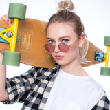 Junge Frau mit blondem Dutt, kariertem Hemd und Skateboard über der Schulter trägt die Hypreader Sonnenbrille Libra Gold/Lila – cooler, urbaner Streetstyle.
