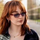 Draußen im Sonnenschein trägt eine junge Frau die Pyxis Sonnenbrille. Die Rot-Violett-Nuancen im Gestell harmonieren mit dem klaren Look der Gläser – ein echtes Statement-Accessoire für Stadt und Freizeit.