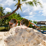 Orangefarbene Libra Sonnenbrille von Hypreader auf einem Felsen am Strand mit Palmen und blauem Himmel im Hintergrund.